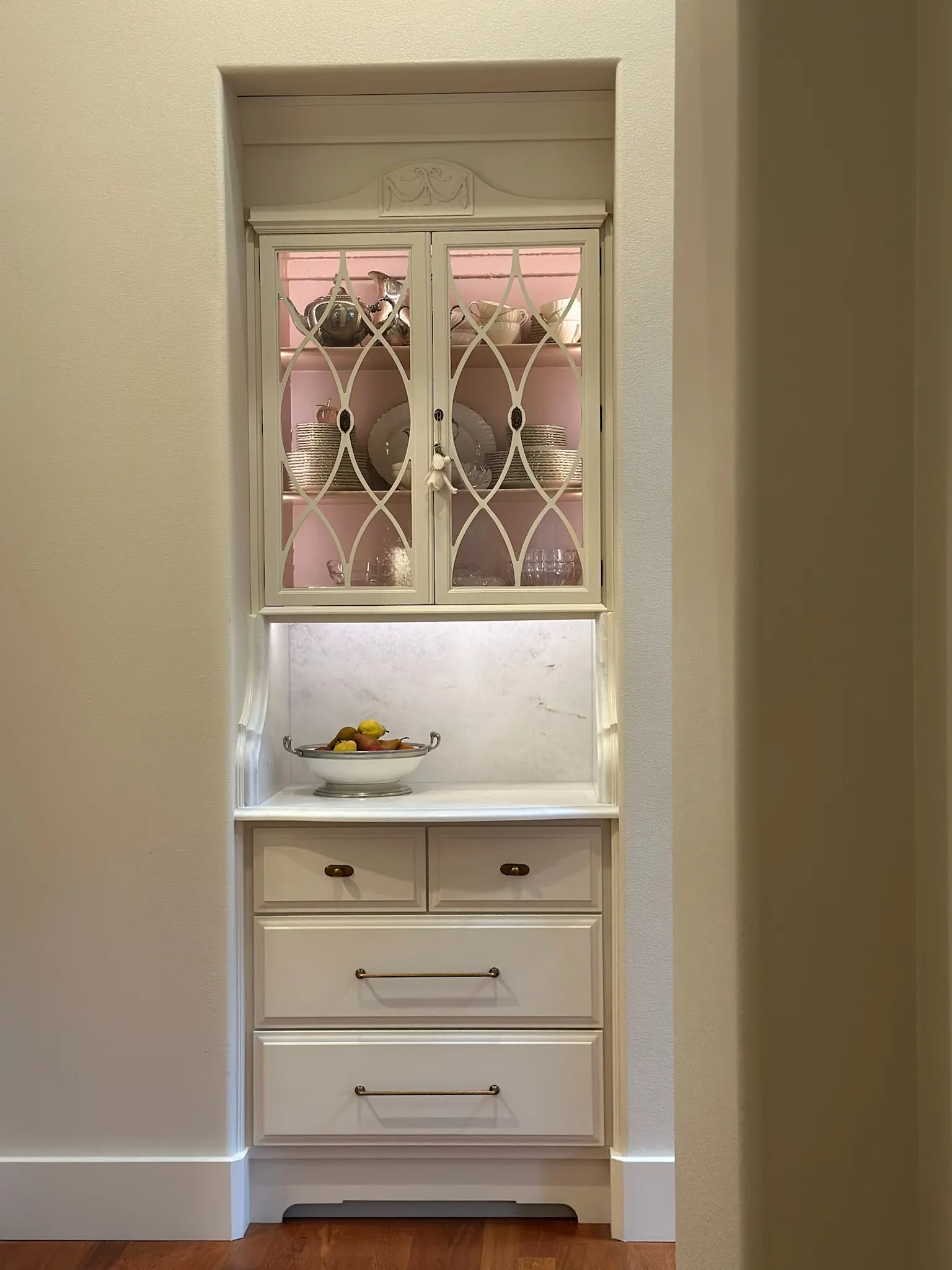 An elegant off-white built-in display cabinet in an alcove, featuring decorative lattice glass doors, pink accent lighting, marble backsplash, and brass drawer pulls.