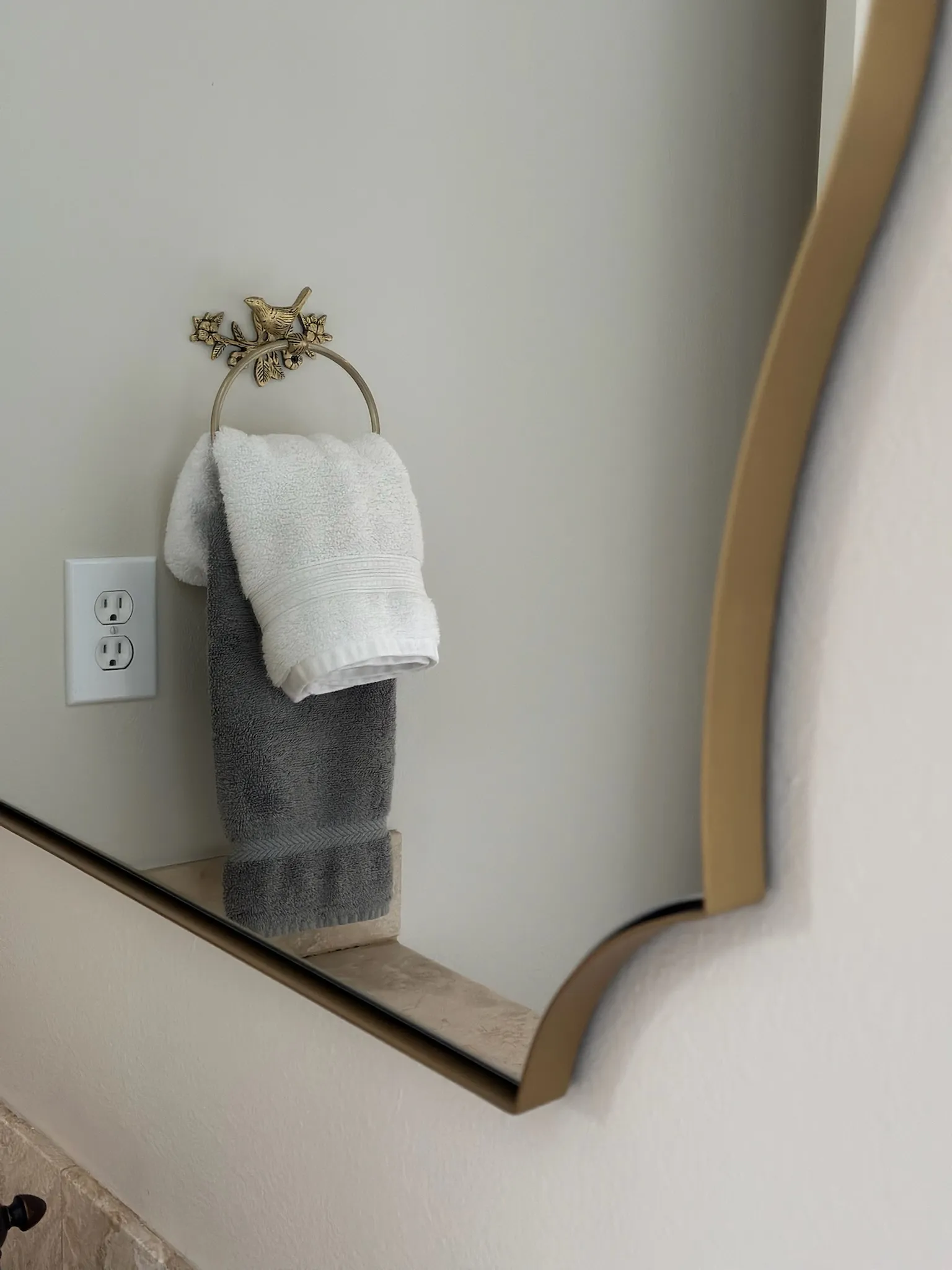 Close-up of a charming brass bird towel ring holding white and grey hand towels in a bathroom, with a golden-framed mirror to the right.
