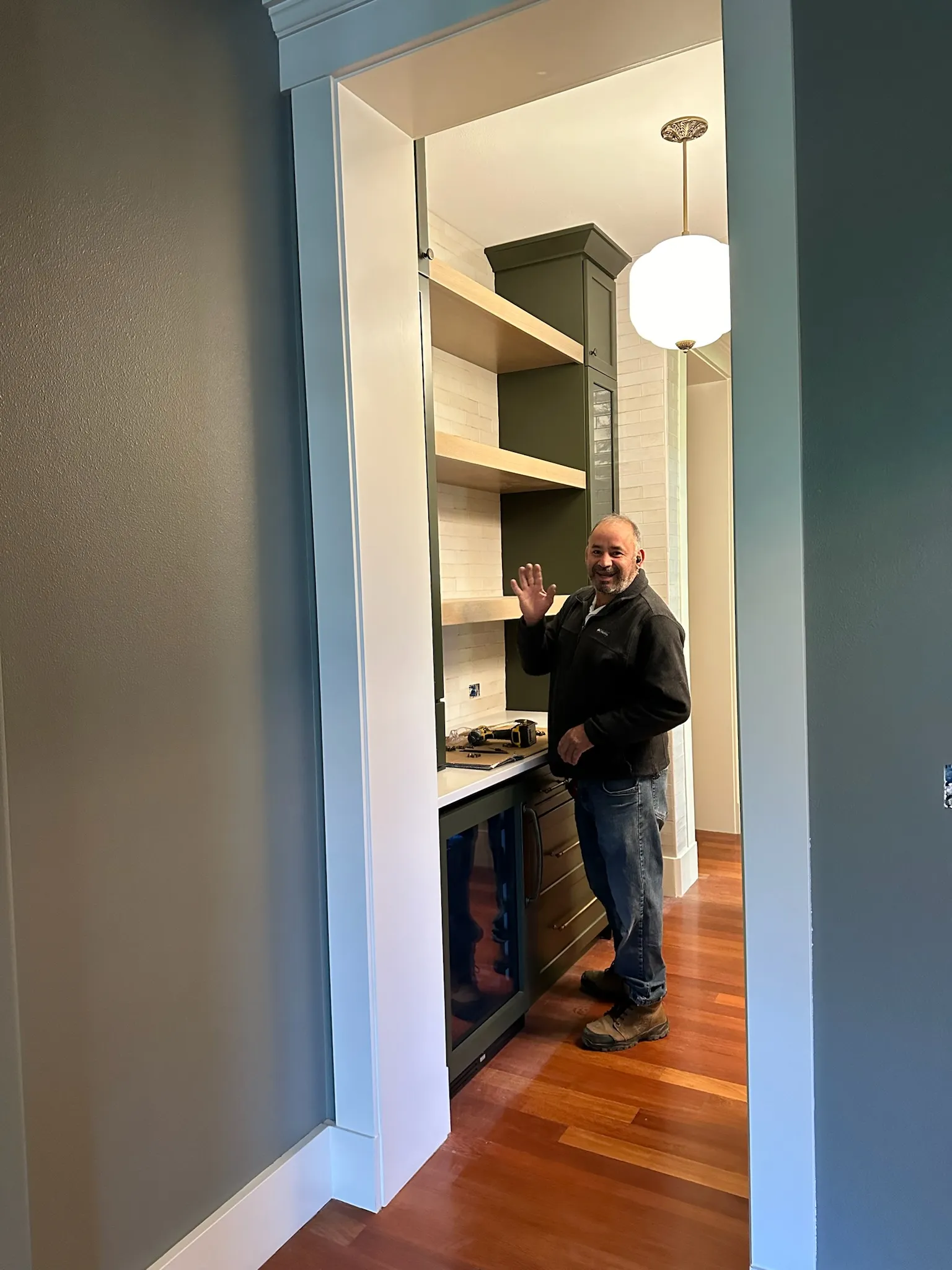 A contractor waves from a newly installed transitional butler's pantry, showcasing dark green cabinetry, light wood shelves, subway tile backsplash, and warm wood floors.