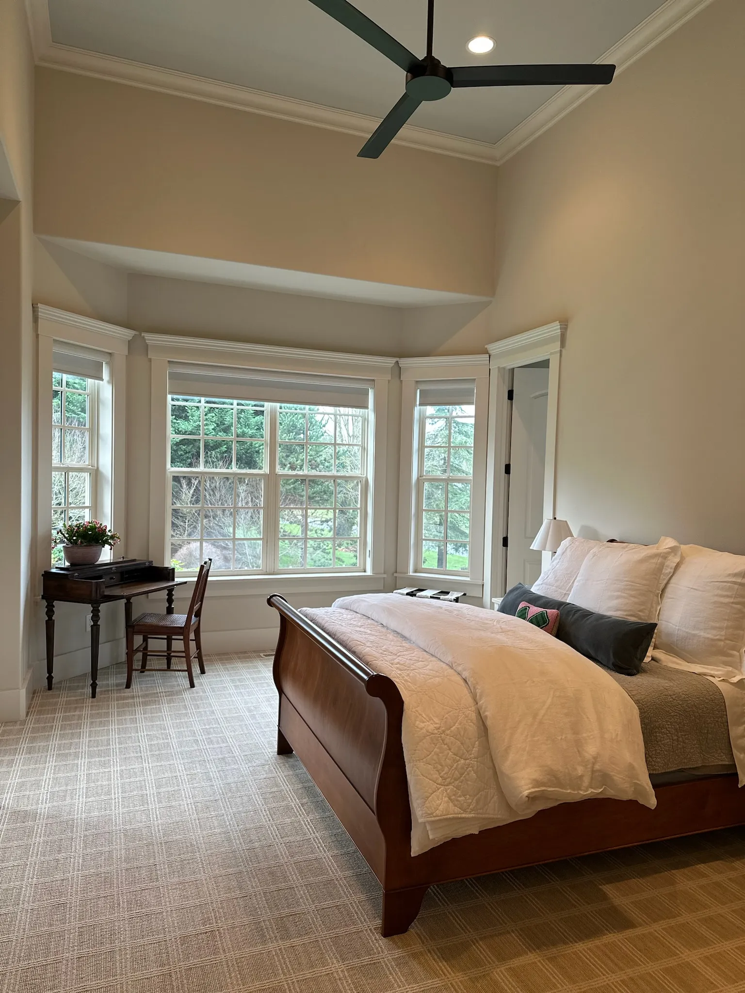 A serene bedroom features a traditional dark wood sleigh bed, bay window with lush views, a writing desk, and patterned carpet. Neutral tones create a calming retreat.
