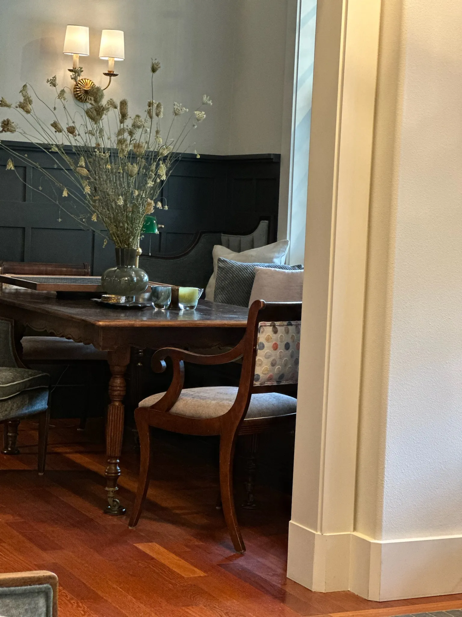 A cozy, eclectic dining room featuring a dark wood table with dried floral centerpiece, ornate wall sconces, and mixed seating against dark paneled walls.