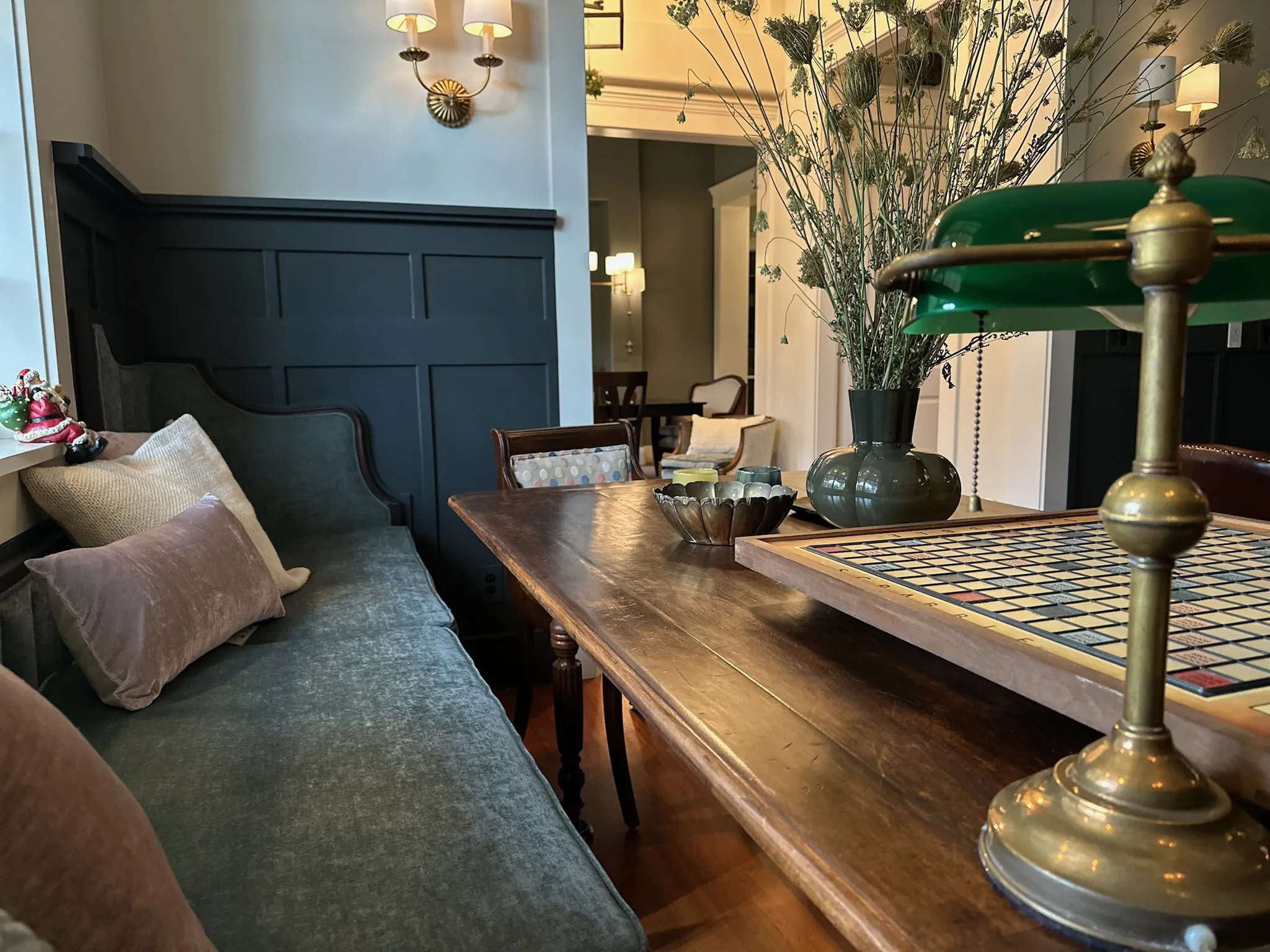 A cozy classic dining nook with a long wooden table, plush green banquette, dark paneled wall, brass accents, and a Scrabble board for inviting leisure.