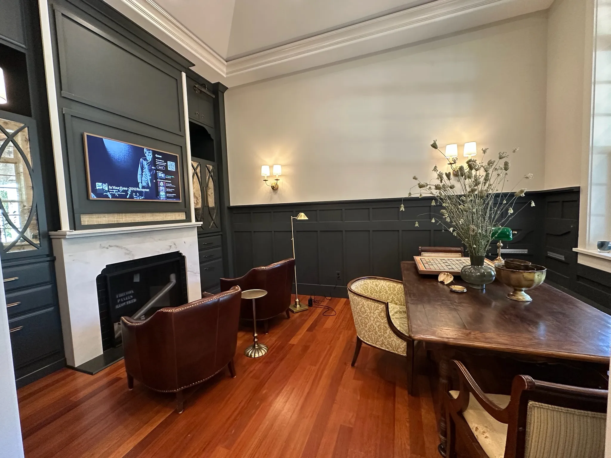 A sophisticated home office featuring dark built-in cabinetry, a fireplace with a TV, a rich wood desk, and classic leather armchairs on warm hardwood floors.
