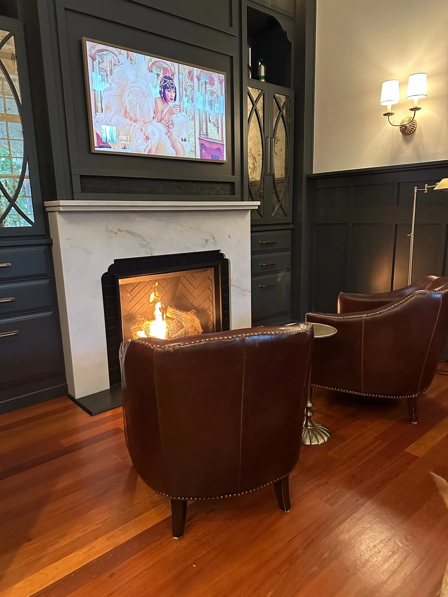 A cozy living room featuring a lit fireplace, flat-screen TV, dark built-in cabinetry, and two classic brown leather armchairs with nailhead trim. Warm and inviting.