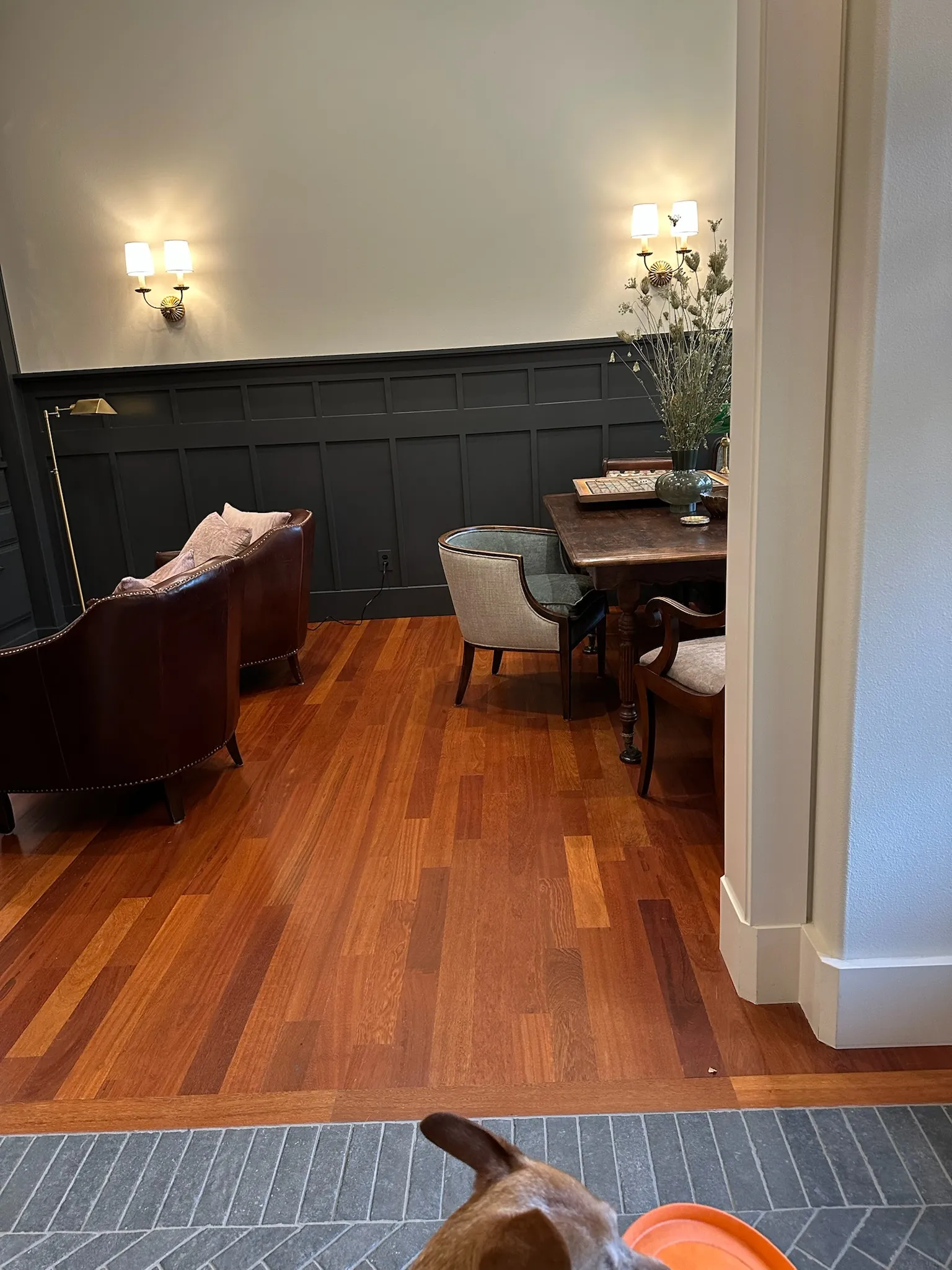 A sophisticated living and dining space featuring dark wood paneling, classic leather armchairs, a rustic dining table, and warm sconce lighting.