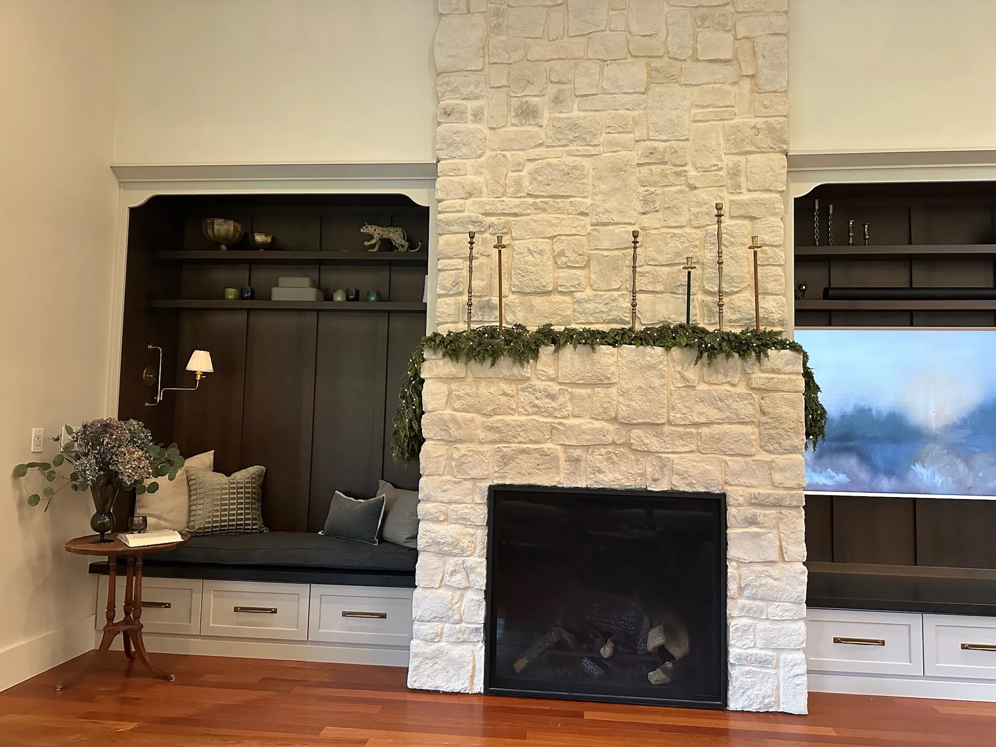 Cozy living room featuring a prominent white stone fireplace adorned with greenery, flanked by dark wood built-ins and a comfortable window seat. Hardwood floors complete the inviting look.
