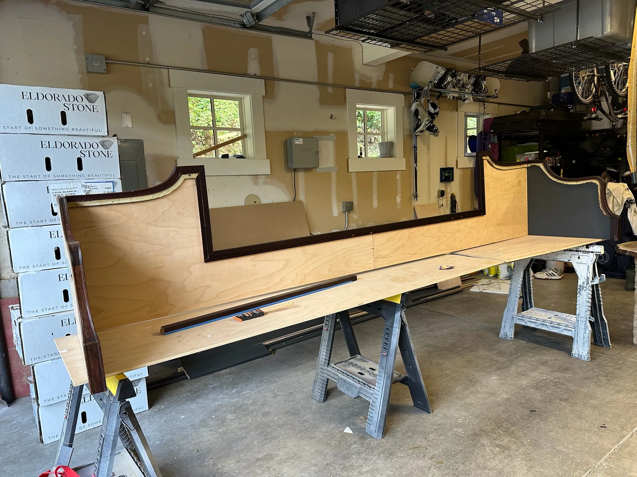 A large custom furniture frame with dark wood trim and a grey panel, under construction on sawhorses in a garage workshop, surrounded by building materials and storage boxes.