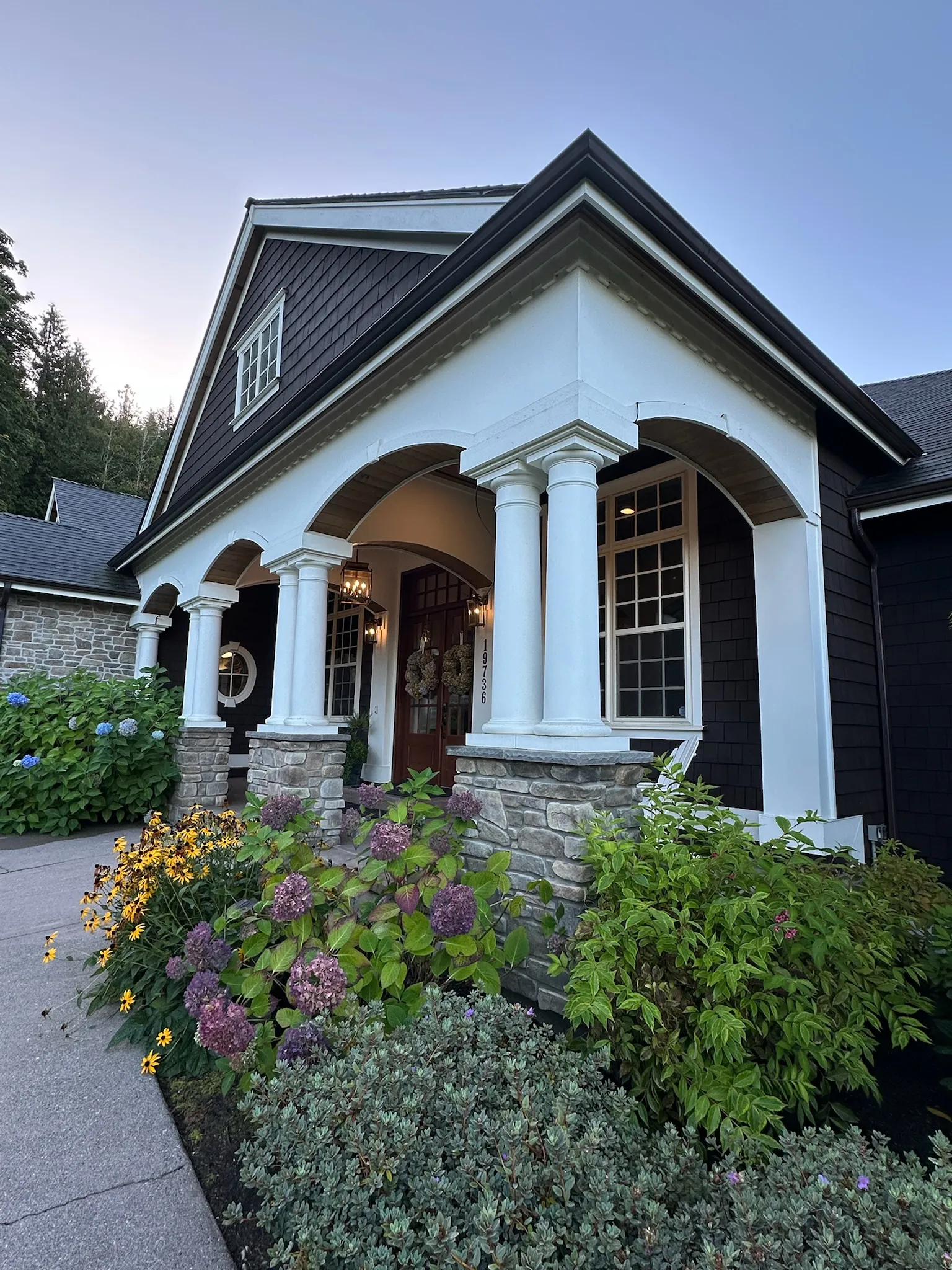 A charming traditional craftsman home exterior with dark shingle siding, white accents, stone columns, and a warmly lit front porch.