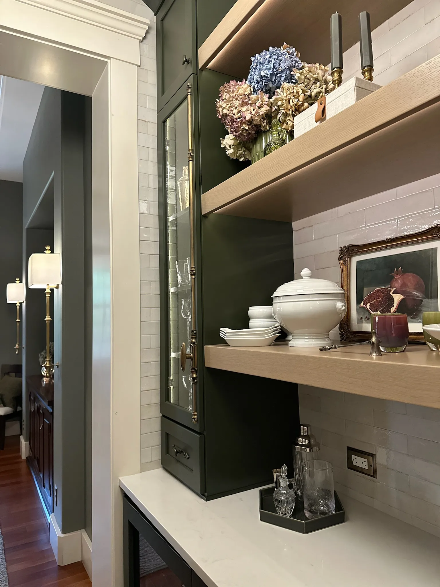 A stylish dark green built-in kitchen shelving unit with open wood shelves, white subway tile, and curated decor, leading to a sophisticated hallway.