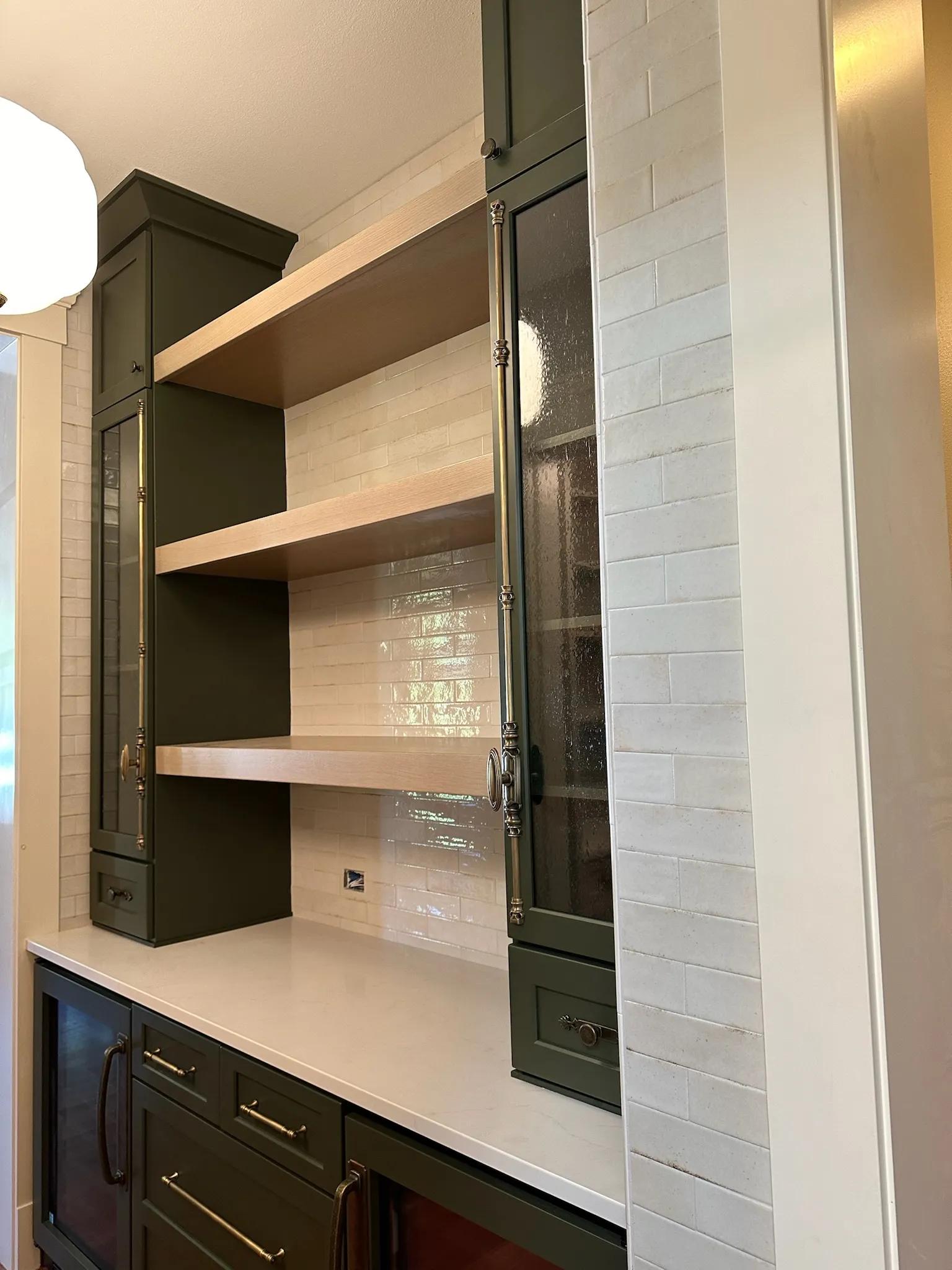 Elegant custom dark green kitchen or bar cabinetry featuring light wood open shelves, a white subway tile backsplash, clean quartz countertop, and sophisticated brass hardware. Perfect for display and storage.