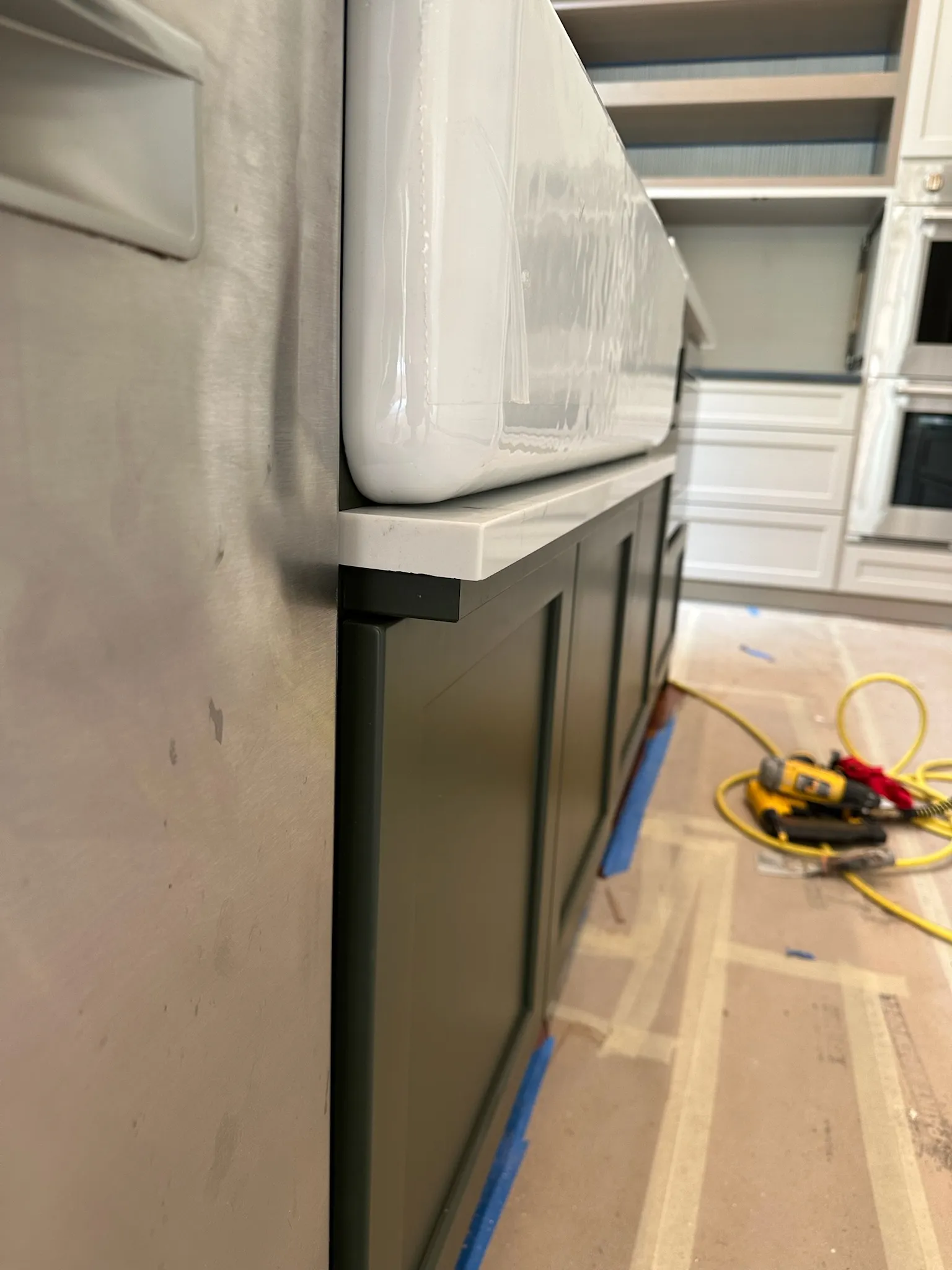 Close-up of a white farmhouse sink being installed on dark green shaker-style kitchen cabinets with white countertops, next to a stainless steel appliance during renovation.