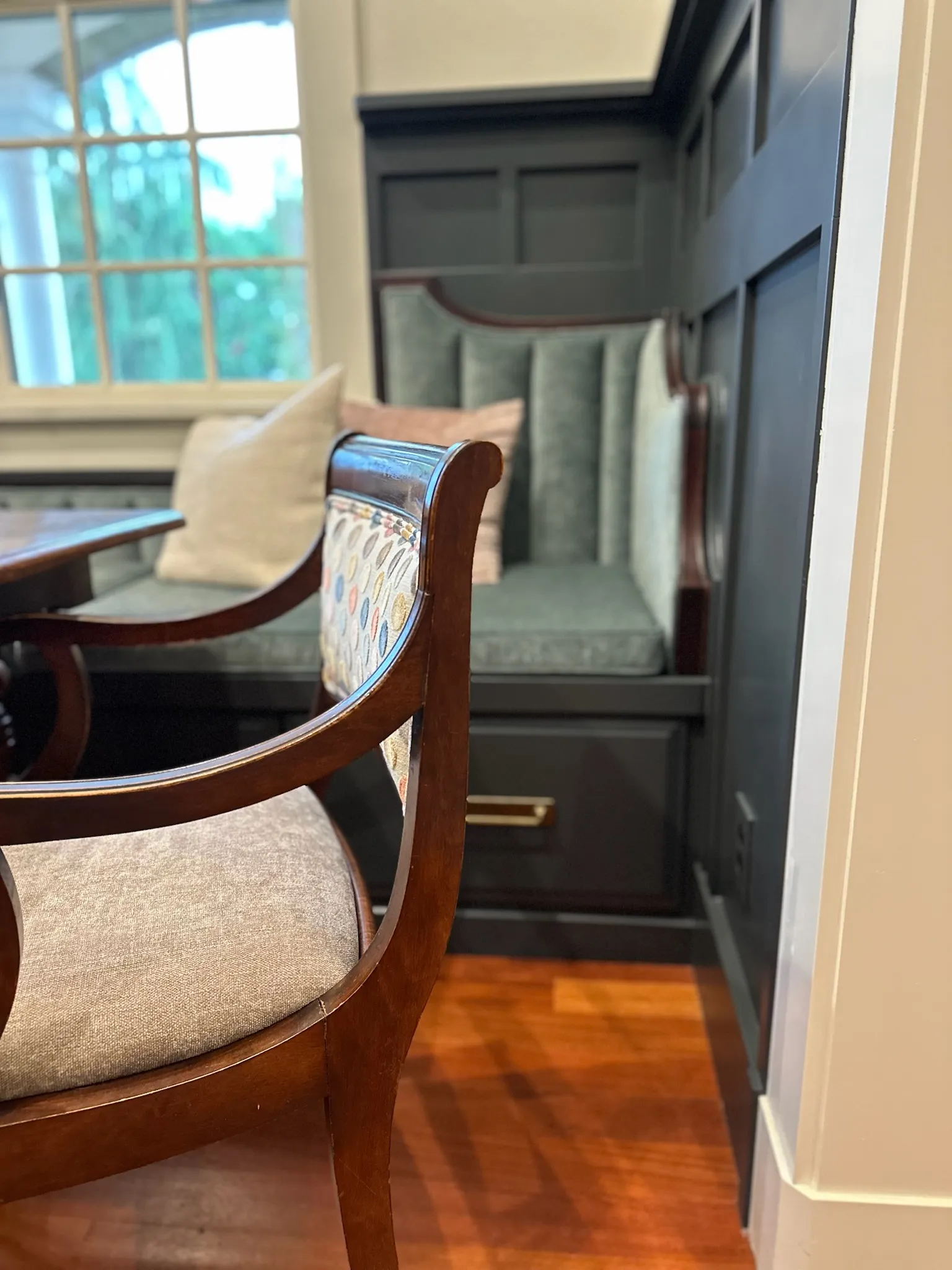 Elegant dining nook featuring a dark built-in banquette with soft cushions, a decorative accent chair, and warm wood flooring bathed in natural light.