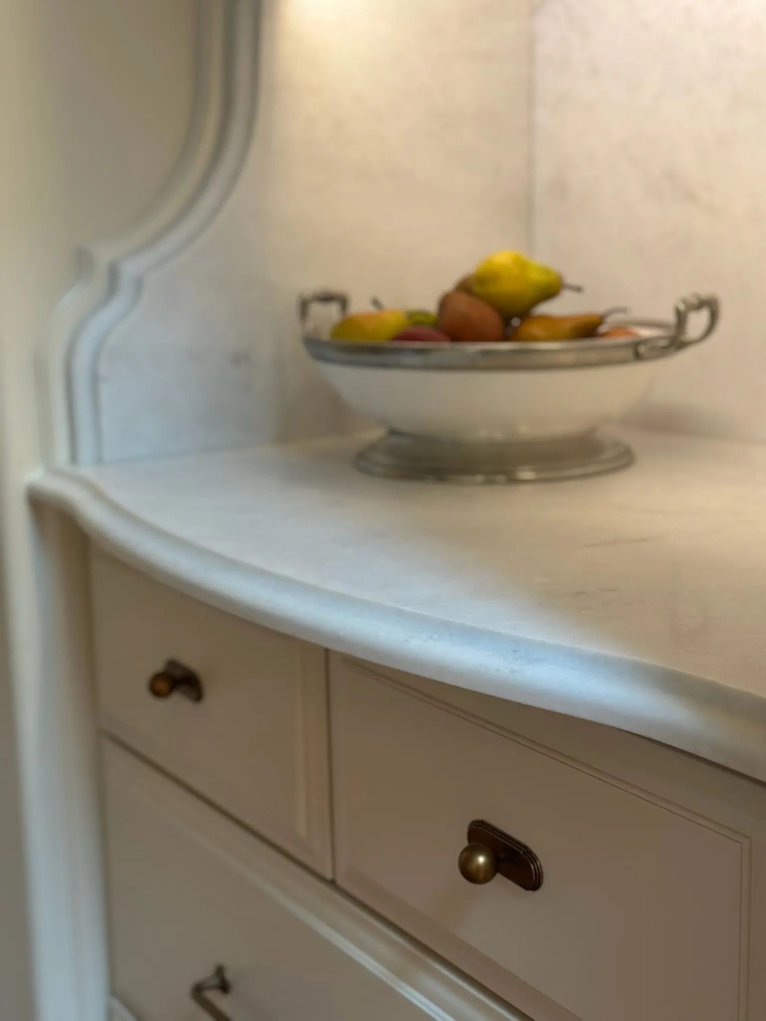 Close-up of elegant light kitchen cabinetry featuring brass pull knobs, a curved marble countertop, and a classic fruit bowl. Blends charm with functional design.