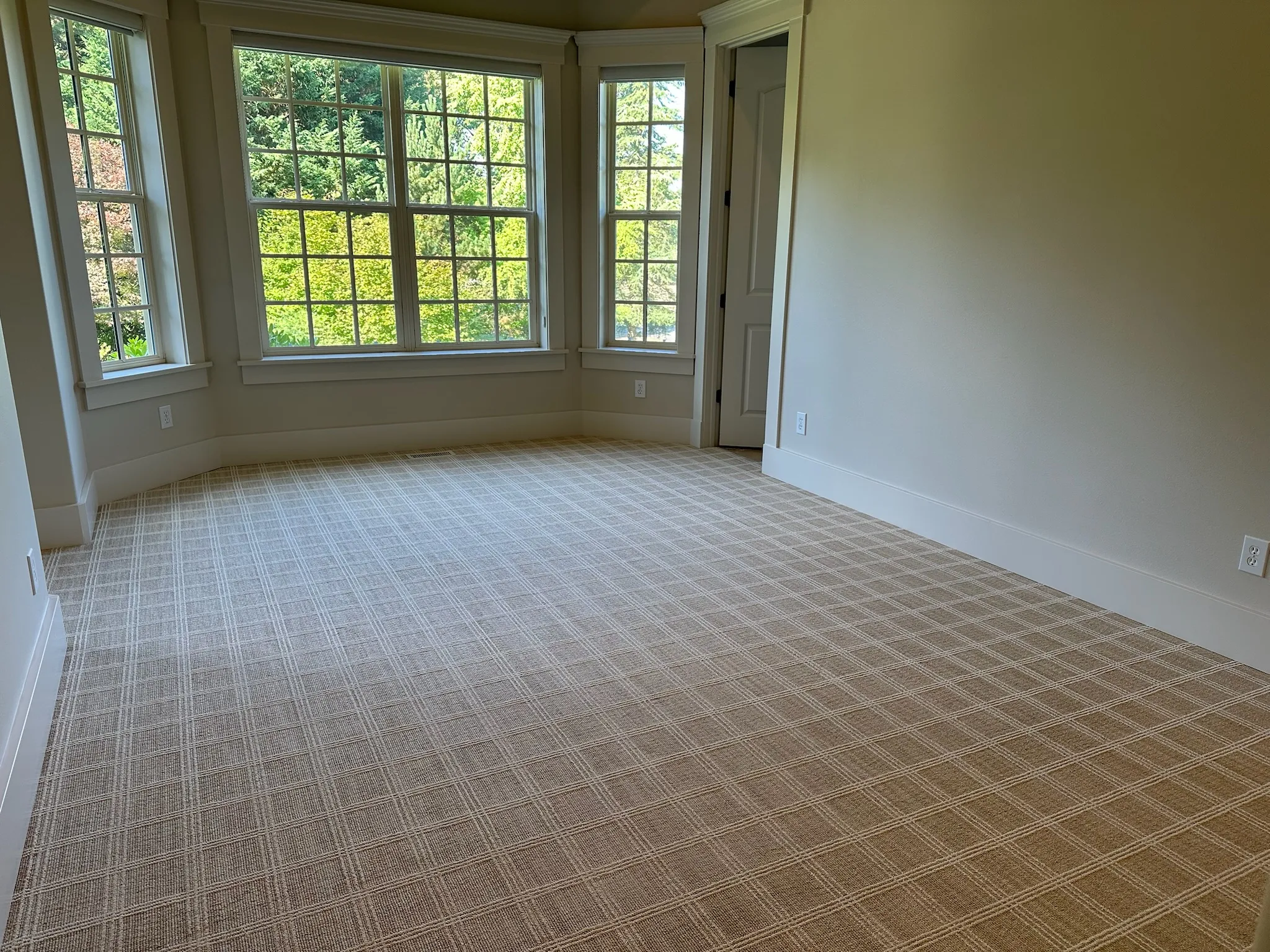 Bright, empty room featuring a large bay window with lush green views, light neutral walls, white trim, and a warm beige plaid patterned carpet. Ready for furnishing.
