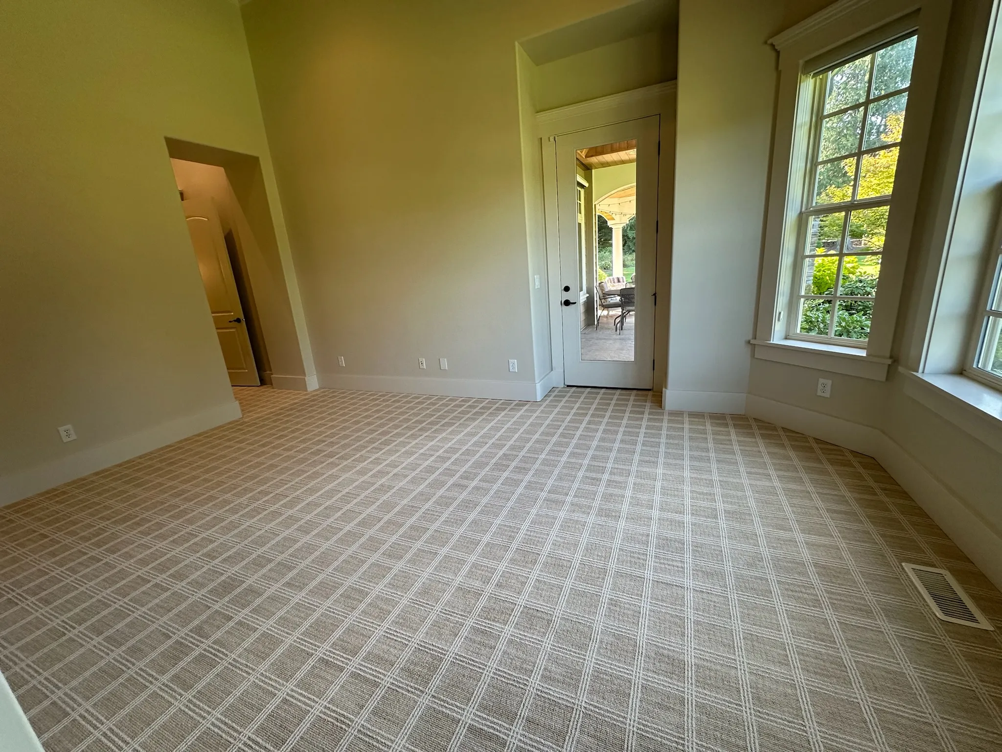 An empty, bright room with light beige walls and a distinctive beige plaid patterned carpet. Natural light streams from a large window and glass patio door, ready for furnishing.