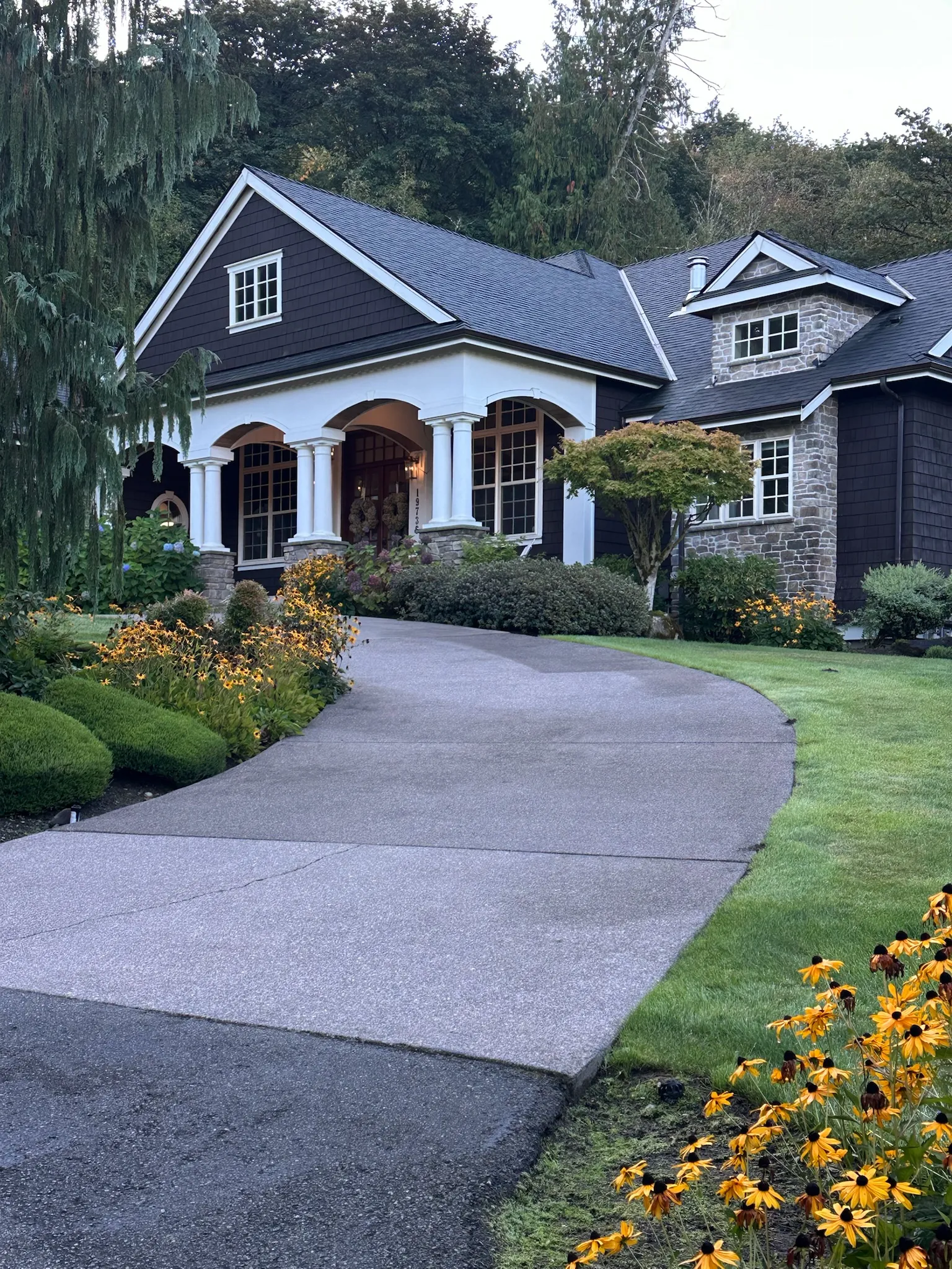 A grand traditional-style home with dark shingle siding, stone accents, and a covered porch framed by white columns, set within lush landscaping.