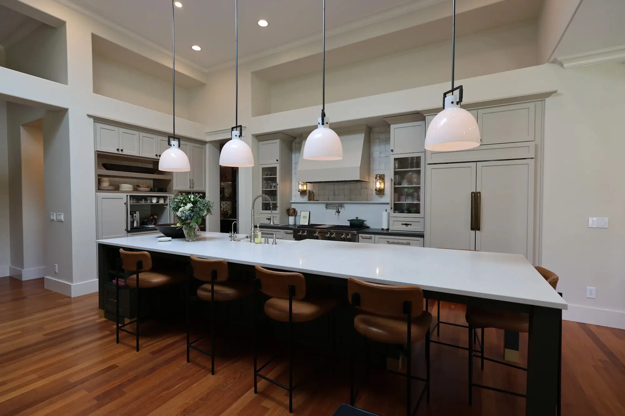 A spacious transitional kitchen featuring a dark green island with white quartz countertop, light neutral cabinetry, and warm hardwood floors. Elegant pendant lights illuminate the space.