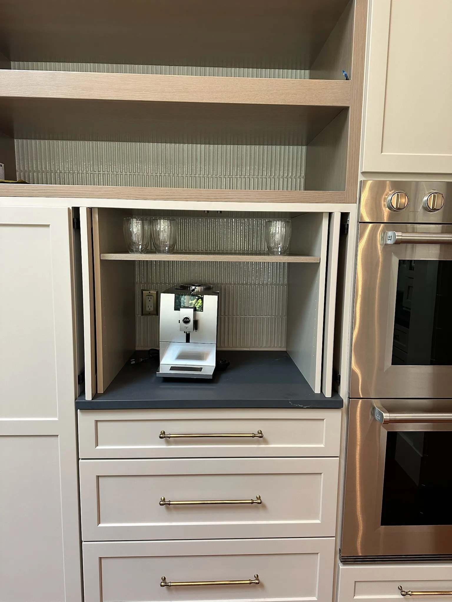 A sleek, built-in coffee bar in a transitional kitchen featuring cream shaker cabinets, brass pulls, a Jura espresso machine, and ribbed tile backsplash.