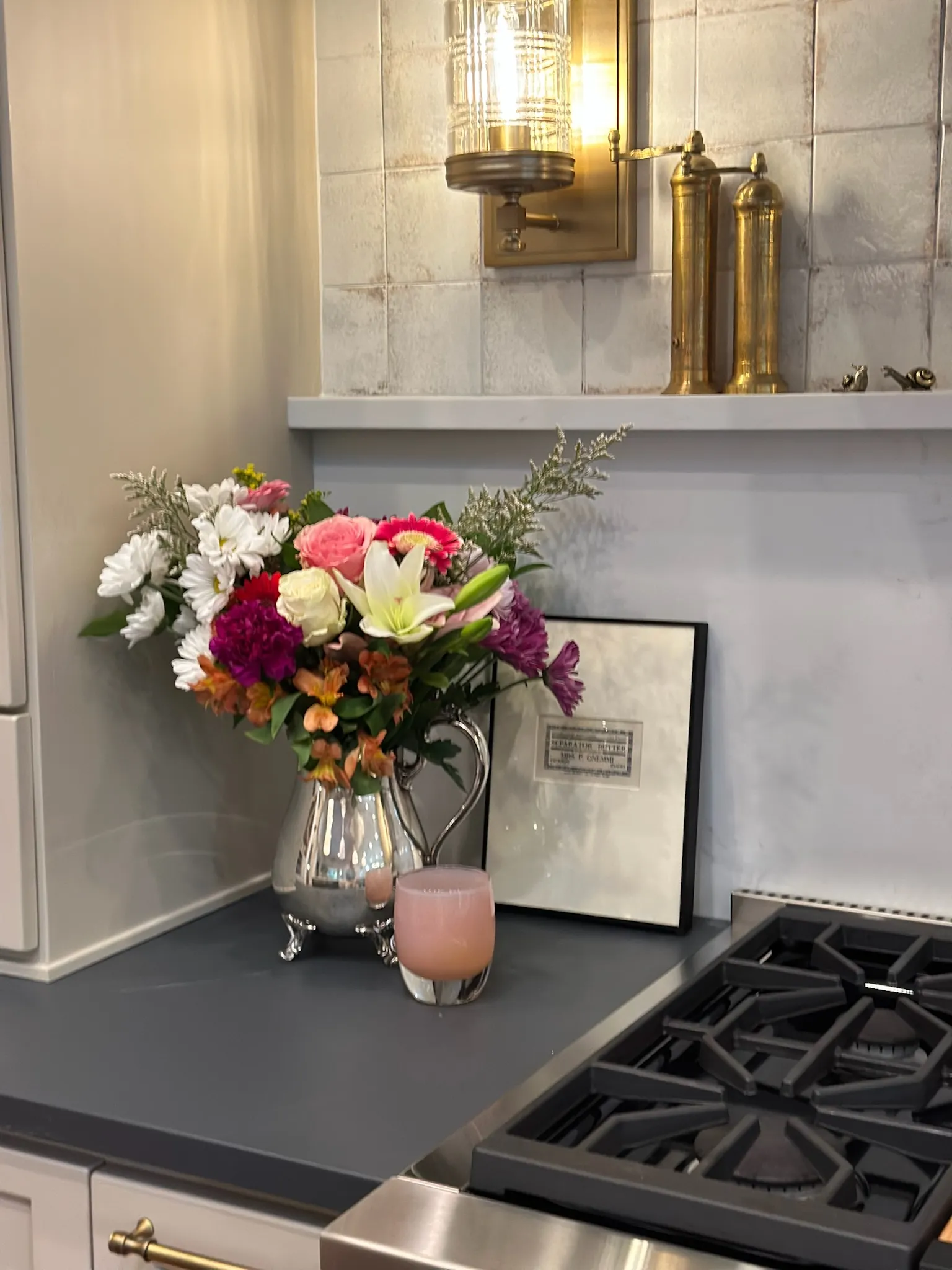 Elegant kitchen counter featuring a vibrant floral bouquet in a silver pitcher, a brass wall sconce, dark grey countertop, and tiled backsplash, exuding warm sophistication.