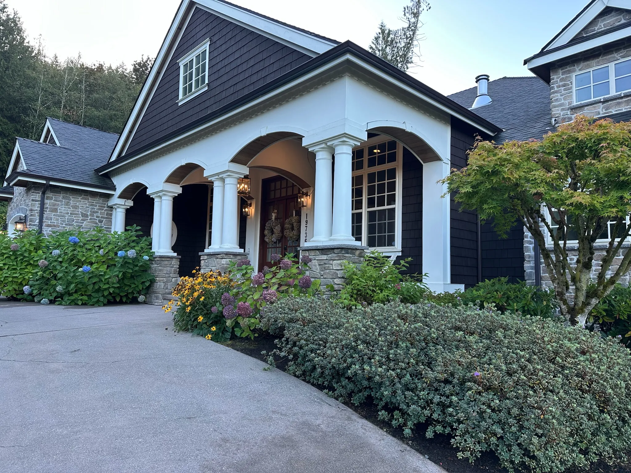 Grand craftsman home exterior featuring dark siding, white pillars, stone accents, and a lush, vibrant garden. A welcoming entryway awaits.
