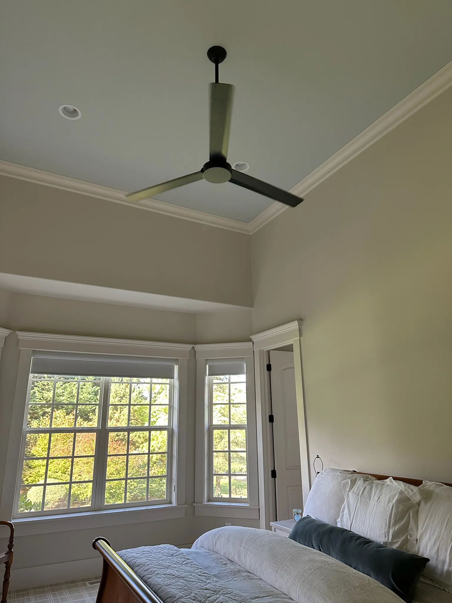 A bright, airy bedroom featuring a modern ceiling fan, large bay windows overlooking lush greenery, and a cozy bed with crisp white linens.