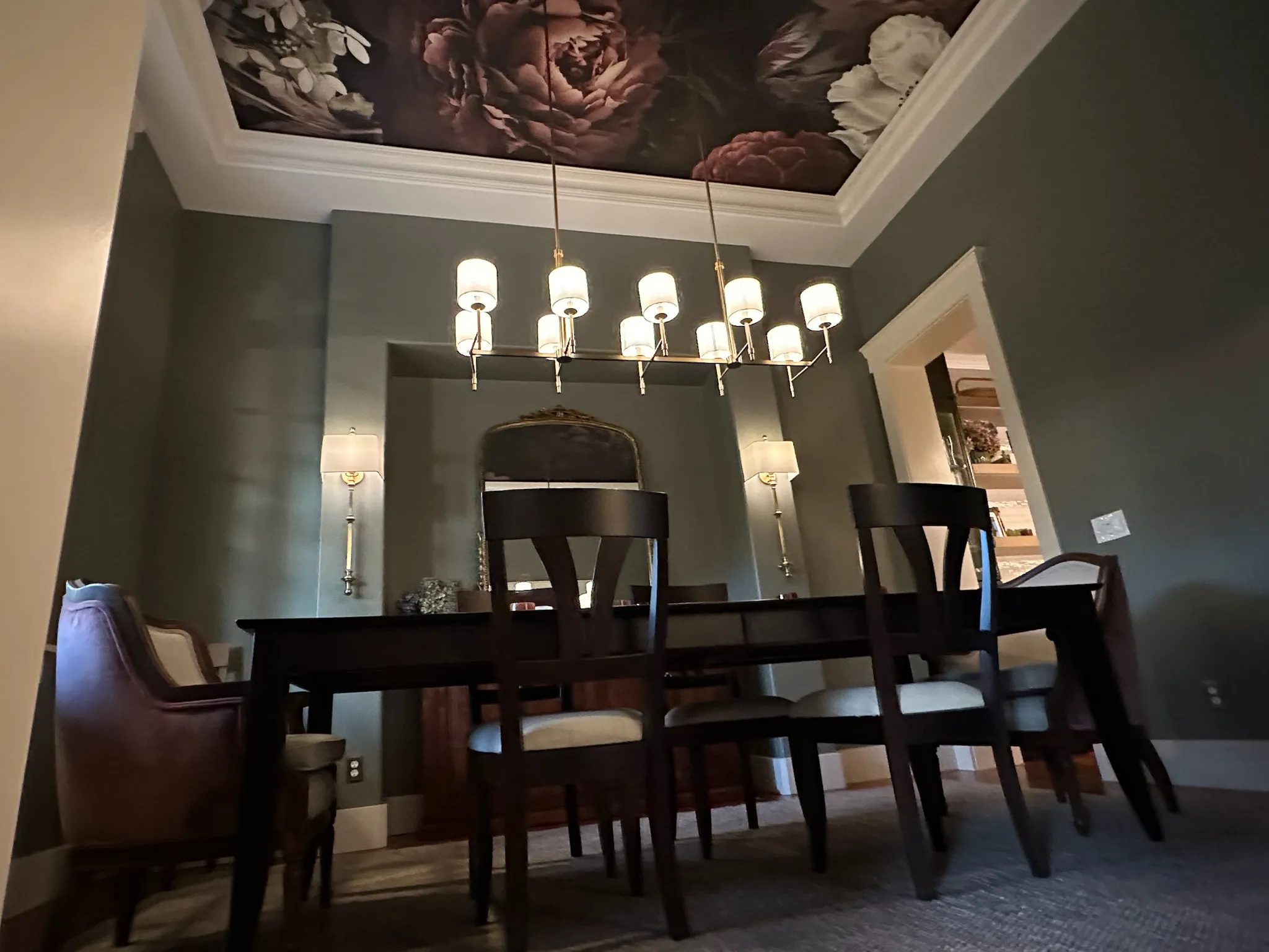 A sophisticated dining room features a dramatic floral ceiling mural, modern linear chandelier, dark dining table with mixed chairs, and deep green walls.