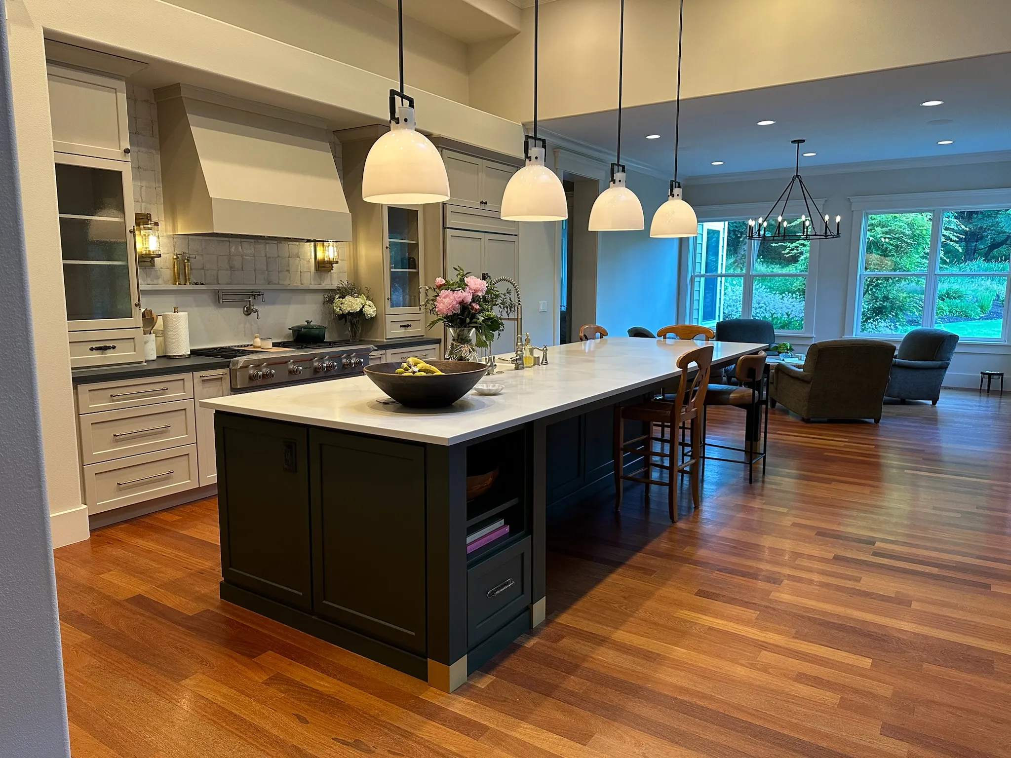 Spacious transitional open-concept kitchen featuring a large dark island with white quartz countertop, modern pendant lights, and light shaker cabinets. Hardwood floors extend into a bright living area with garden views.