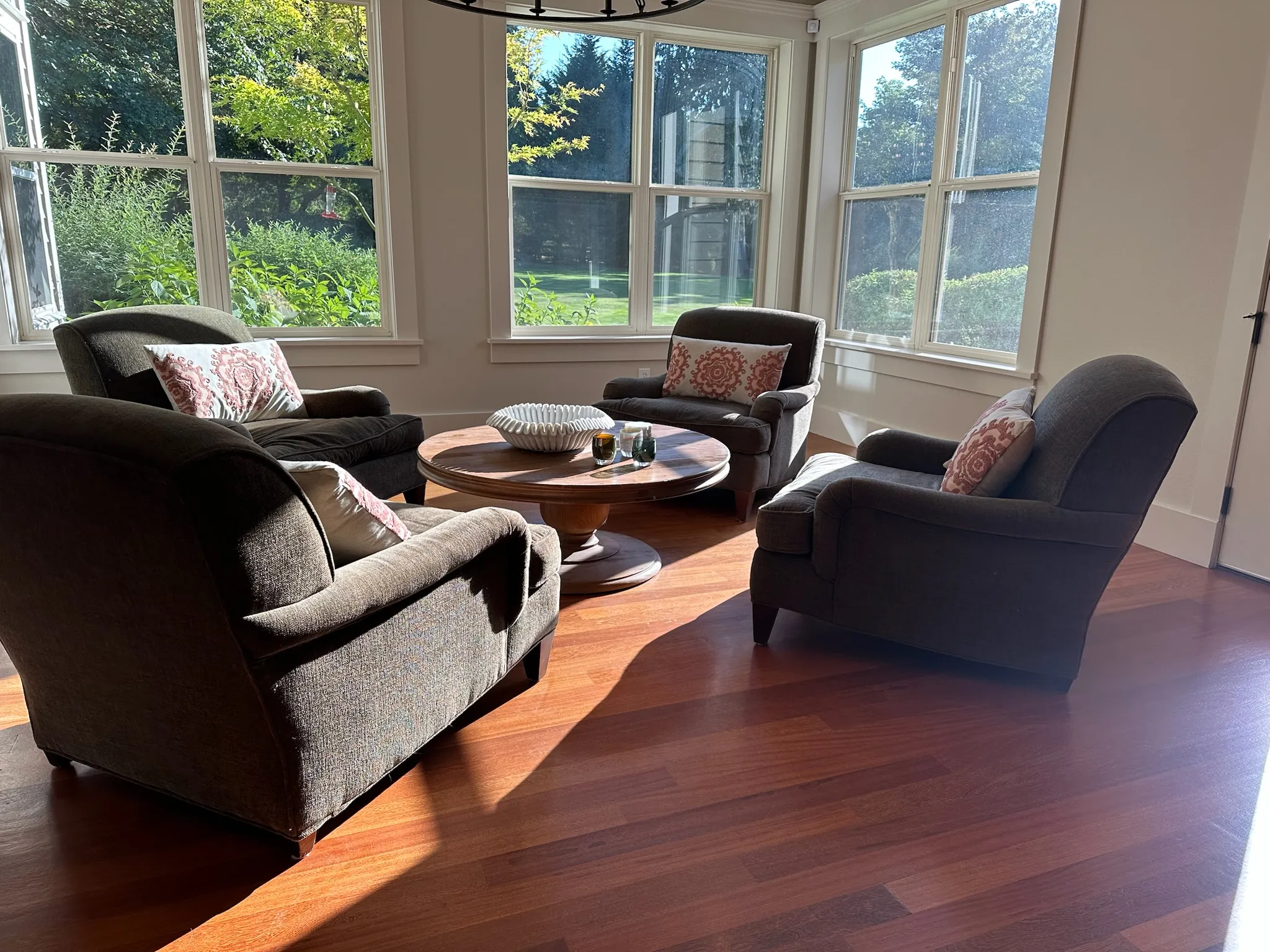 A sunlit living room with four cozy, dark brown armchairs arranged around a round wooden coffee table, offering a serene view of lush greenery outside.
