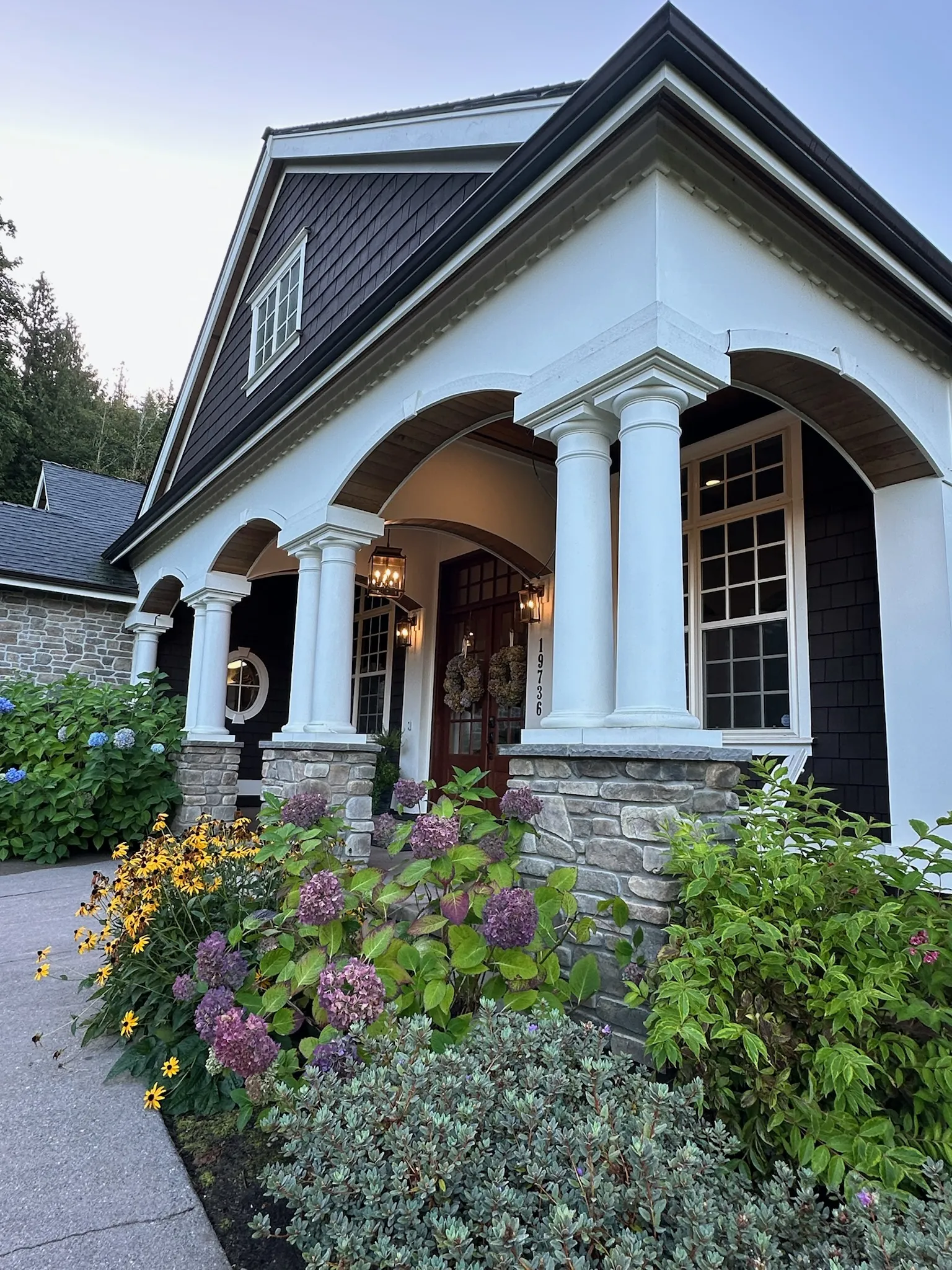 An inviting traditional craftsman home entryway featuring dark shingle siding, white columns, stone details, and vibrant landscaping leading to the front door.