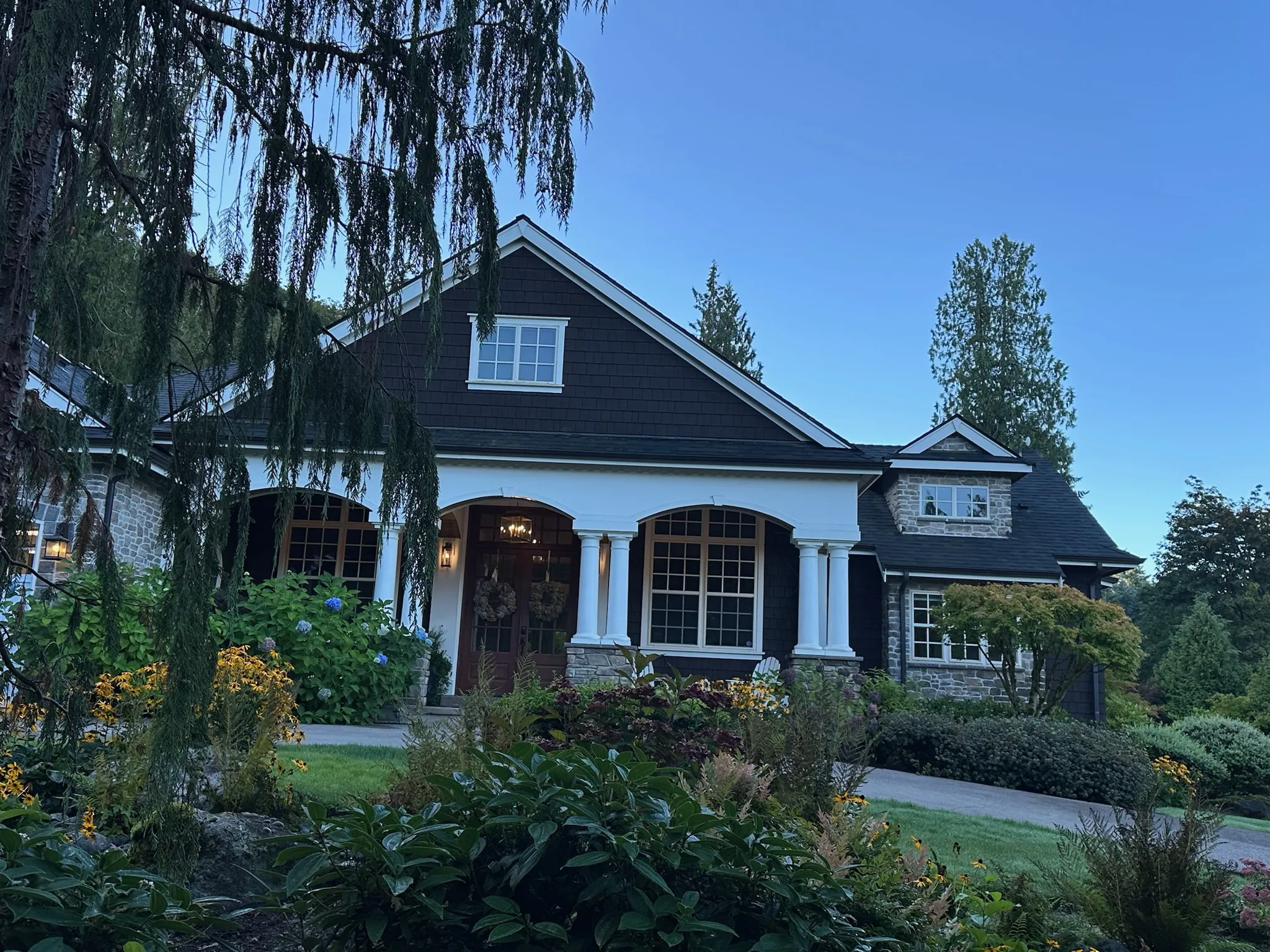 Elegant Craftsman-style home with dark shingle siding, stone accents, and a welcoming white-columned porch, surrounded by lush, mature landscaping at dusk.