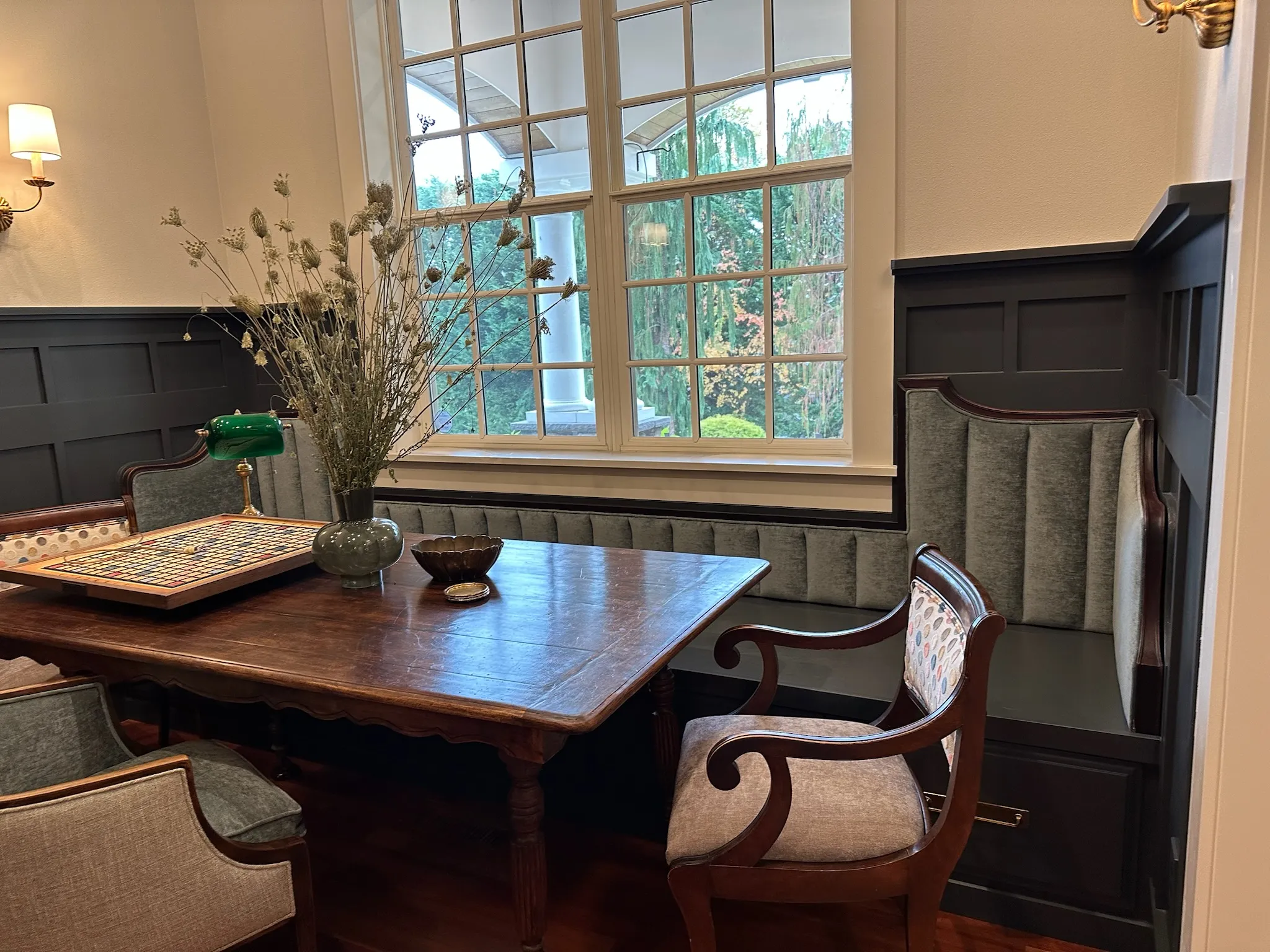 A sophisticated traditional dining nook featuring a dark wood table, elegant upholstered chairs, and a custom built-in banquette with fluted gray upholstery, illuminated by a large window.