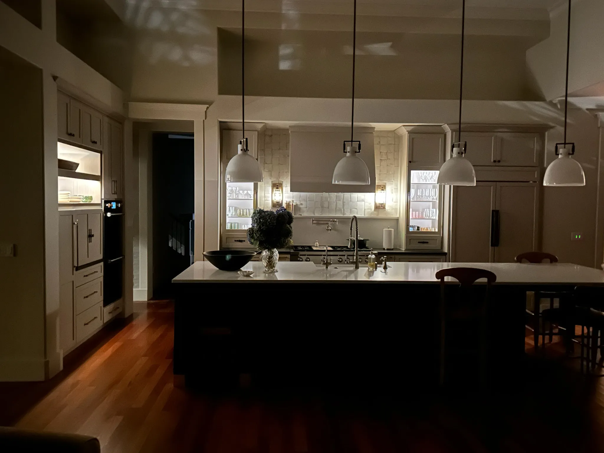 Spacious transitional kitchen with a large island, sleek white pendant lights, warm wood floors, and modern built-in appliances. Cozy evening ambiance.