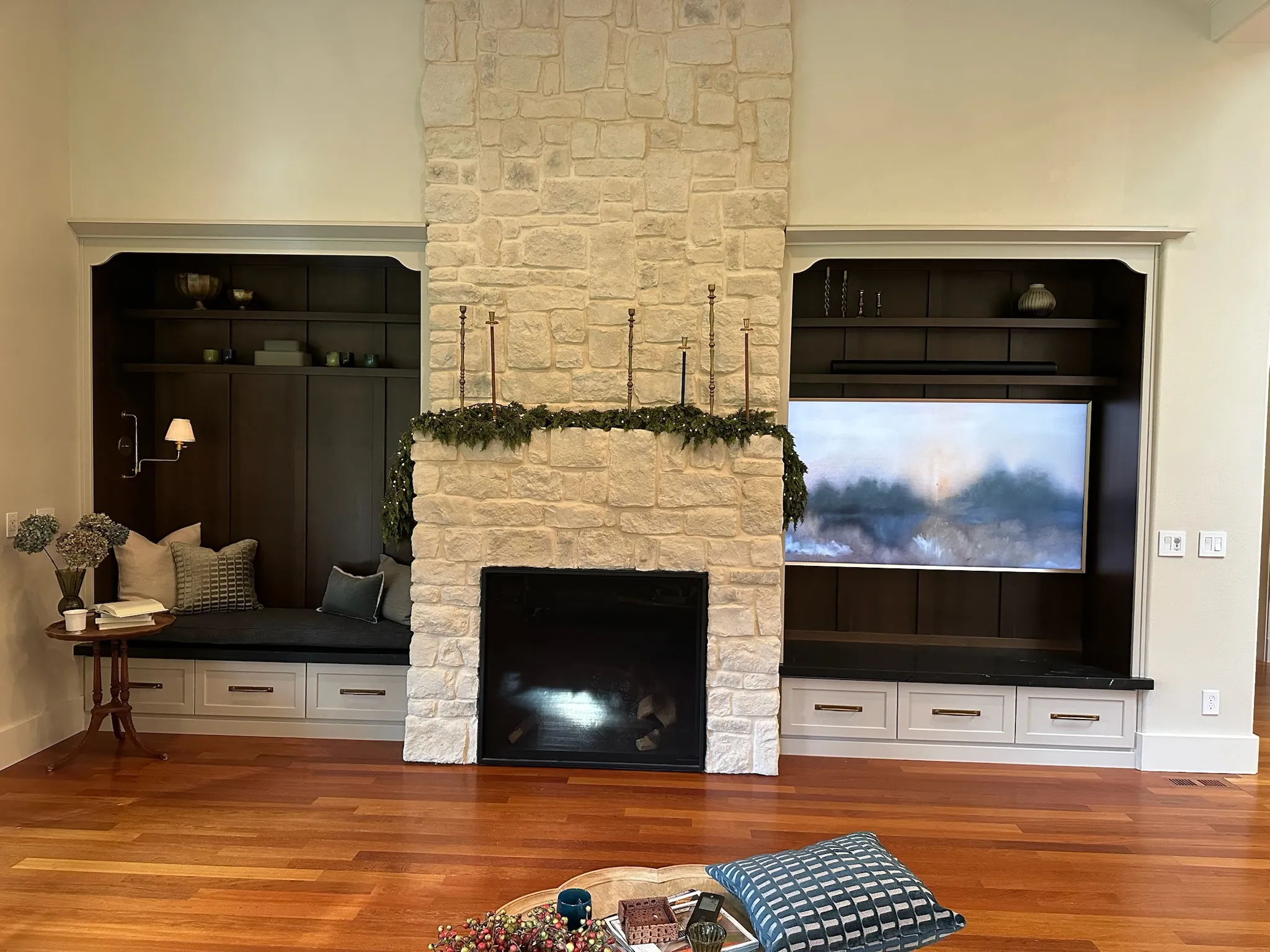 A warm and inviting living room featuring a grand stone fireplace flanked by custom dark wood built-in shelving, a cozy window seat, and an integrated TV.