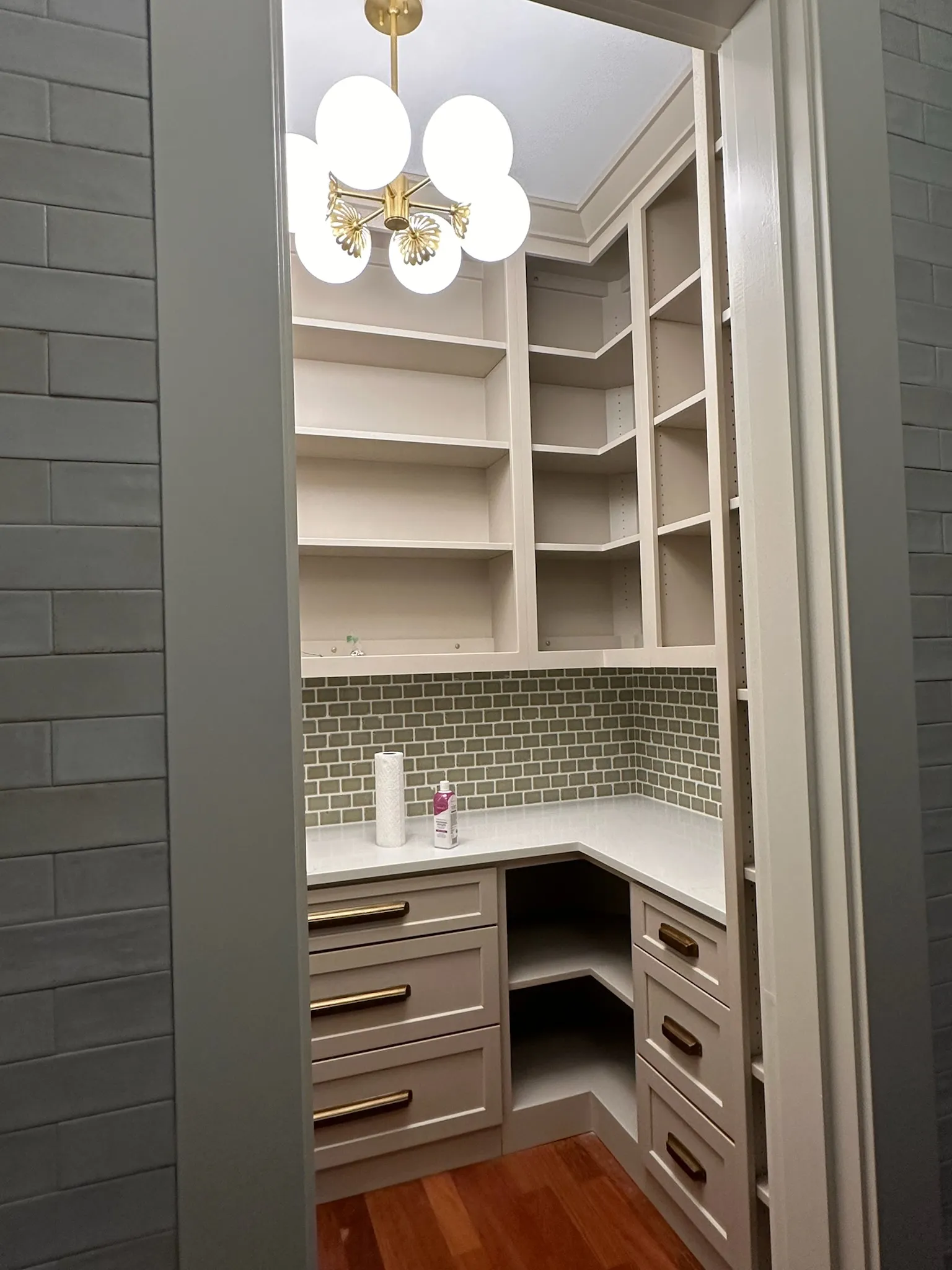A stylish transitional walk-in pantry featuring light beige custom cabinetry, integrated shelving, brass hardware, green subway tile backsplash, and a modern globe chandelier.